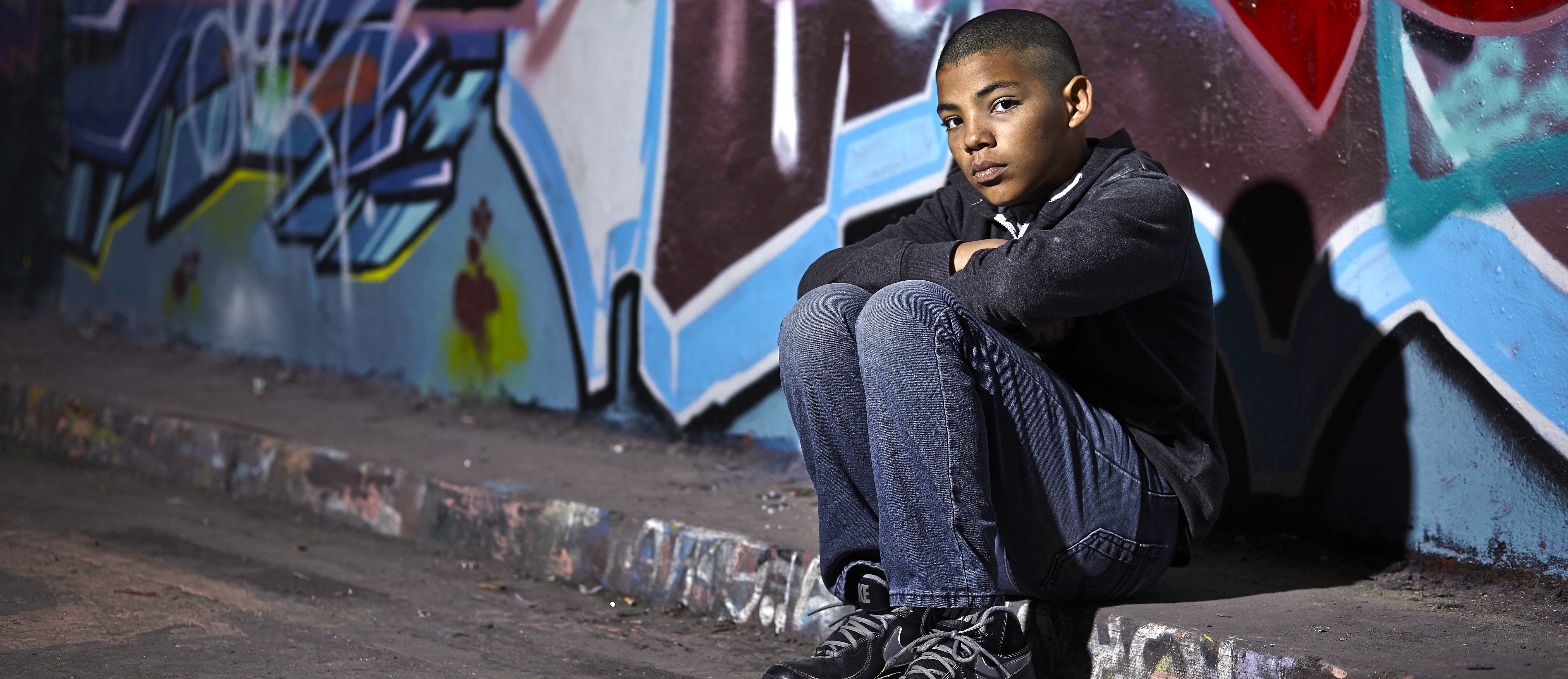 Boy sat on the floor of an alleyway in front of a graffiti wall