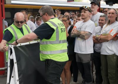 Volunteers wearing high-vis jackets setting up a stand next to Three Peaks By Rail participants wearing white T-shirts and standing on a train station platform before the event.