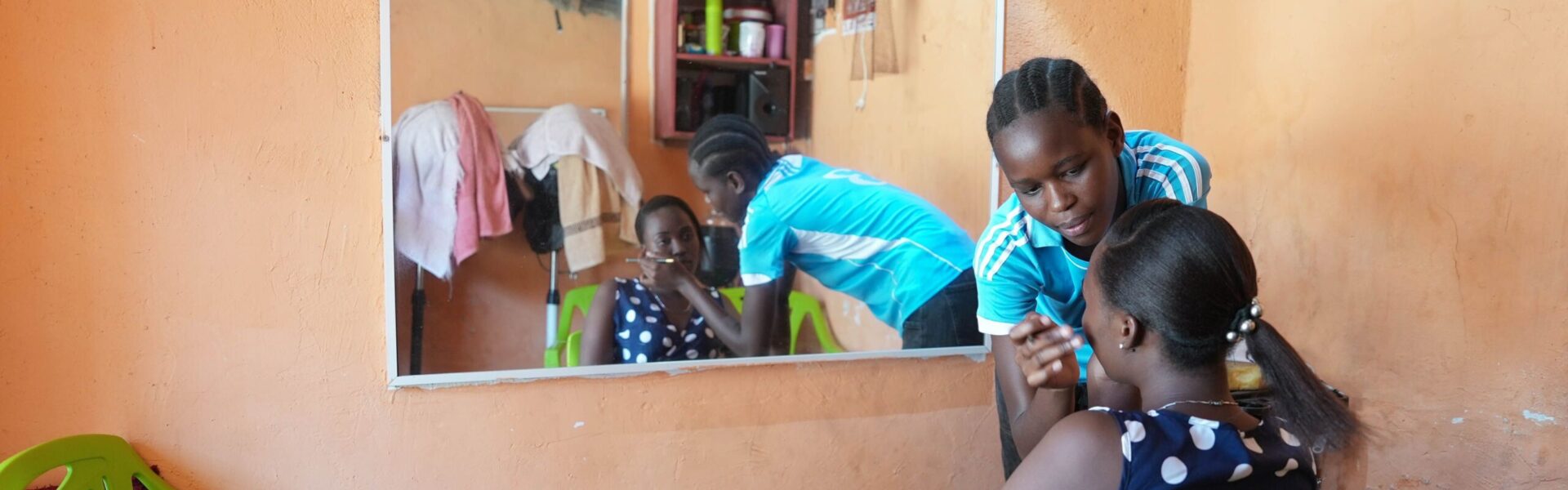 A young woman in a blue shirt applies makeup to another woman seated on a green chair in a small room with orange walls. Their reflection is visible in a large wall mirror behind them.
