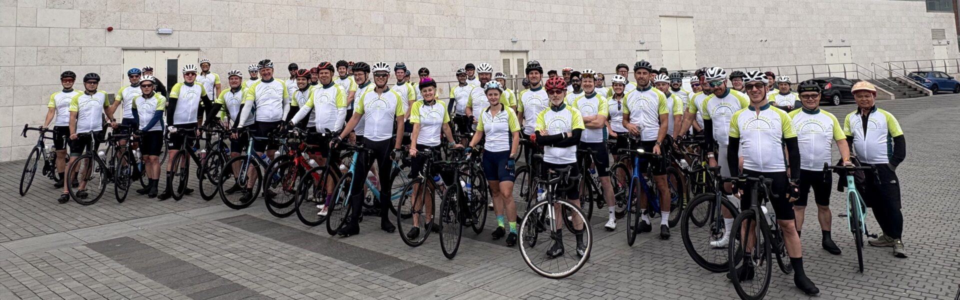 A group of Ride the Route participants posing on bikes in front of a modern building and wearing green and white branded Ride the Route / Railway Children cycling jerseys.