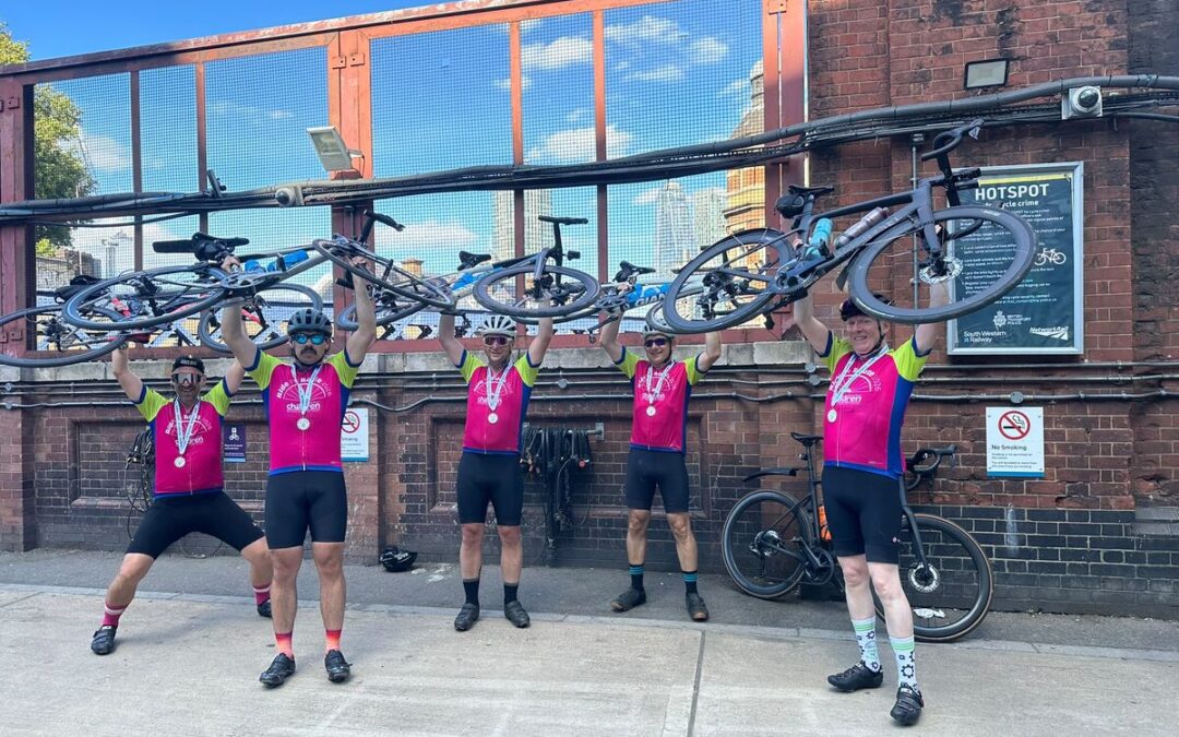 Five cyclists wearing brightly coloured clothing holding up their bikes above their heads in a row.