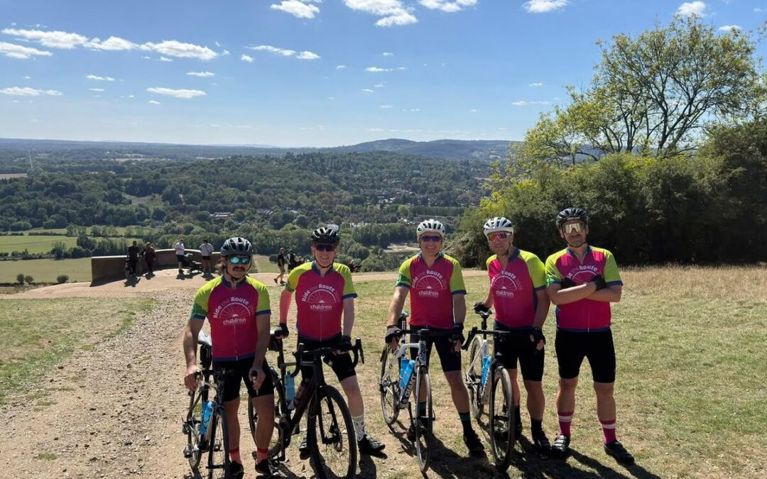 Five Ride the Route cyclists pose with their bikes at a scenic hilltop viewpoint overlooking a valley on a clear sunny day.