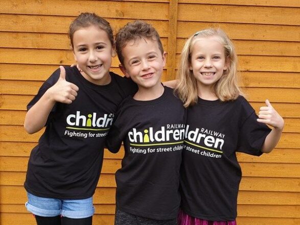 Three young children wearing black railway children t-shirts - with their thumbs up for the camera.