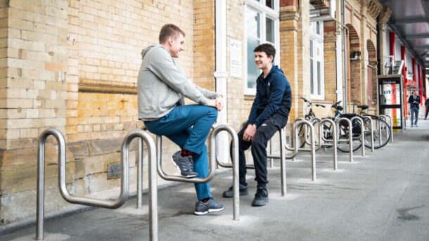 Two teenage boys sat talking to each other at a train station.