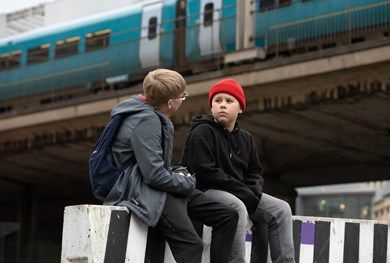 Two teenagers sat below a rail bridge, a blue train passing by in background.