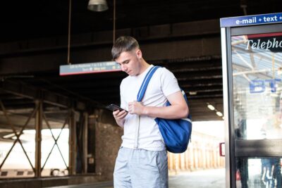 A teenage boy standing at a station platform, scrolling on social media on his phone and carrying a blue backpack.