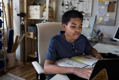 Teen boy sat at a desk with a laptop and book.