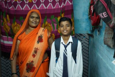 A mother wearing orange stood with her son who is in a school uniform at their home in India.
