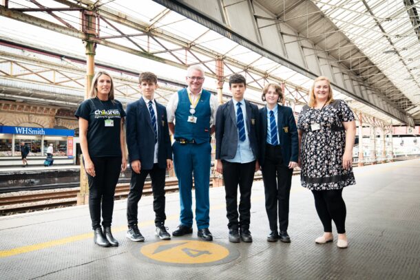 A Railway Children youth practitioner, three young teenagers, an Avanti West Coast staff member and a social worker all stood in a group on a train station platform in the UK.