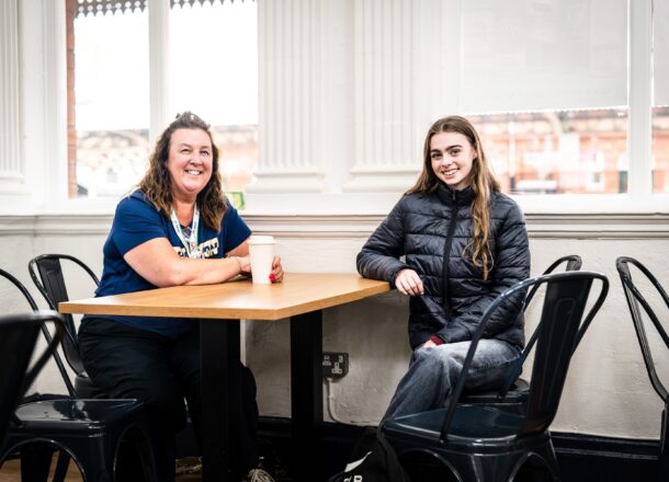 A Railway Children youth practitioner sat in a cafe with a young person at a table, both are leaning on the table and smiling.