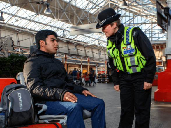 A British Transport Police officer talking to a teenage boy who is sat on a bench at a train station in the UK with a backpack next to him.