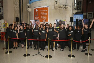 A group of people at a Railway Children Corporate event - The Midland Pullman. They are wearing Railway Children T-shirts and standing behind a red rope at Paddington Station in London.