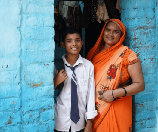 Indian boy in school uniform and mother in orange sari stand in doorway smiling at camera