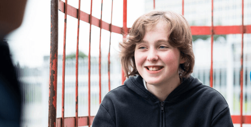 A teenager sat on a trolley outside a train station, smiling and looking into the distance.