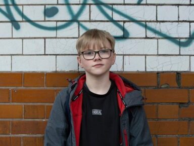 Young boy with glasses stood against brick wall.