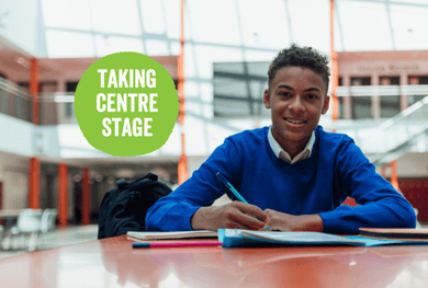 Teen boy in blue school jumper sat at a desk with a pen in his hand. Text in a green circle reads: Taking centre stage.