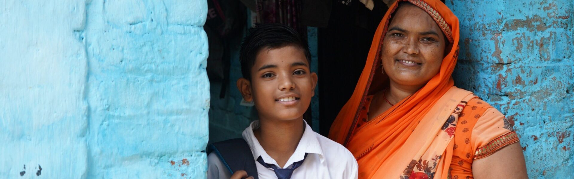 Indian boy in school uniform and mother in orange sari stand in doorway smiling at camera