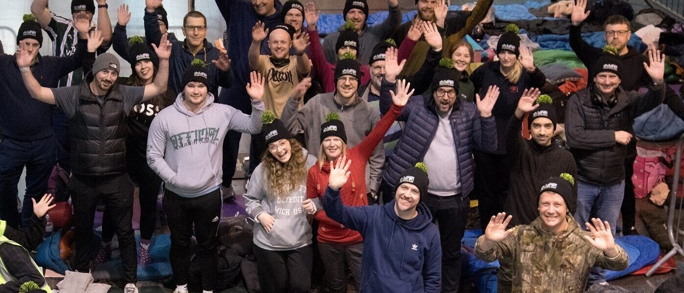 A group of people taking part in Railway Children's Sleepout event at Manchester Piccadilly wearing branded black bobble hats and waving at the camera.