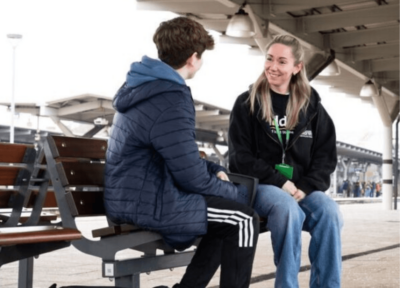 Blonde youth practitioner worker smiling talking to child with blue coat on train platform.