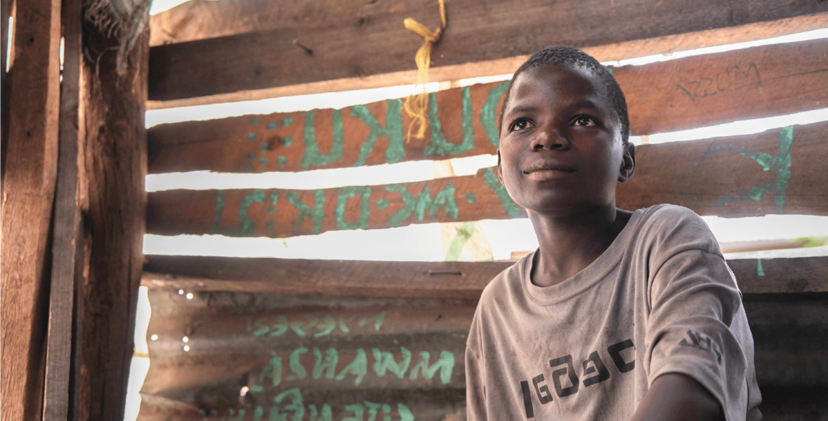 A boy sat in front of a wooden shed on a bench in Tanzania.