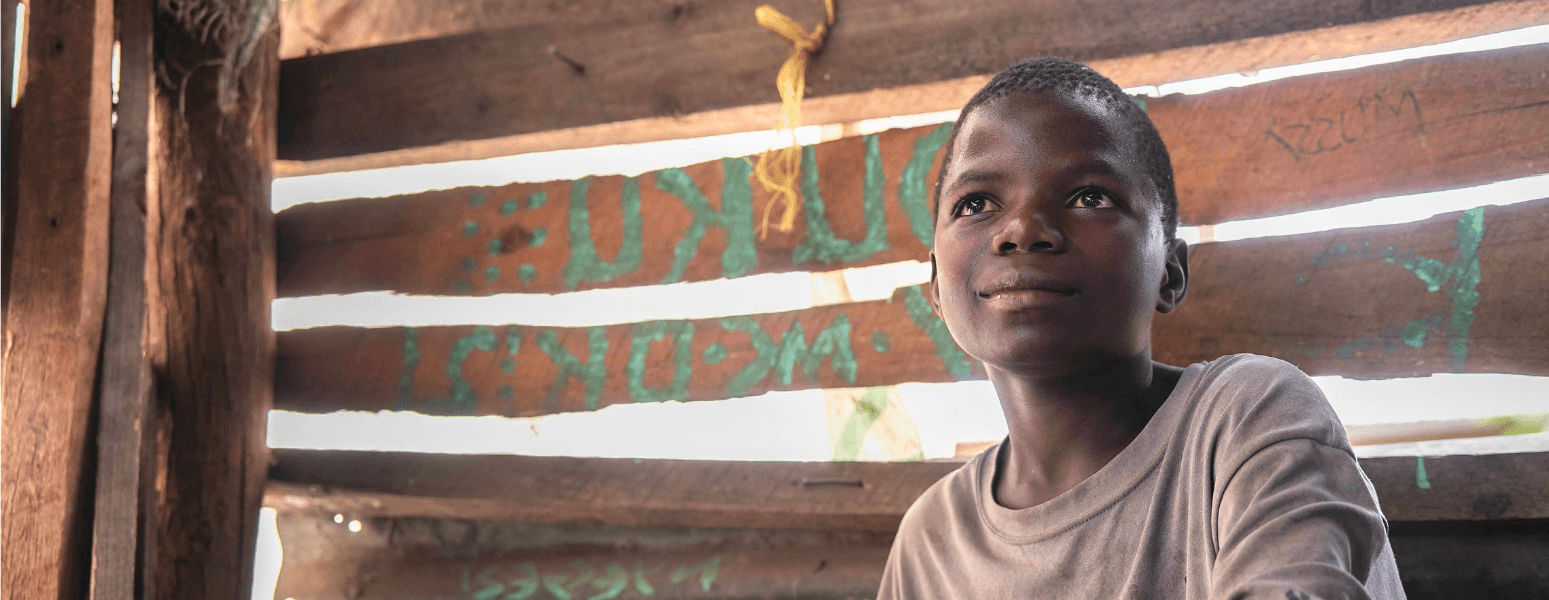 A boy sat in front of a wooden shed on a bench in Tanzania.