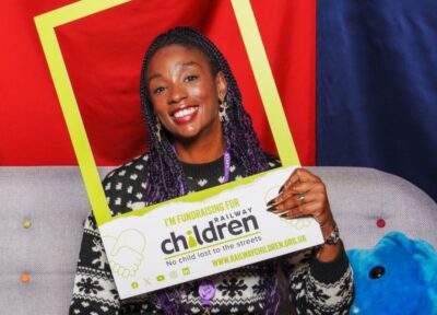 A woman wearing a Christmas jumper, sitting on a sofa and holding a Railway Children selfie frame which says on it 'I'm fundraising for Railway Children'.