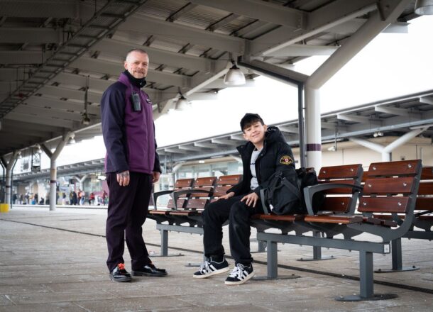 A male member of staff working for the rail network wearing a purple jacket and dark trousers, stood with a teenage boy who is sitting on a bench at a train station.
