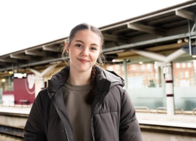 A teenage girl wearing a brown puffer jacket stood on a railway station platform in the UK.
