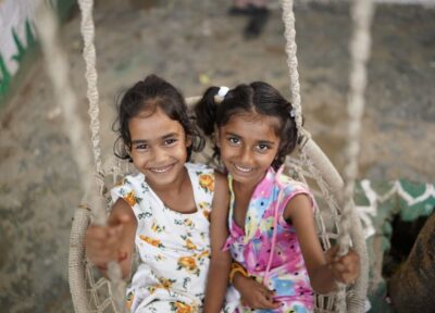 Two young girls sat in a swing together. Both smiling for the camera.