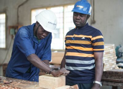 Two men wearing safety helmets in a woodworking workshop, one instructing the other on carpentry techniques while working with a wooden box