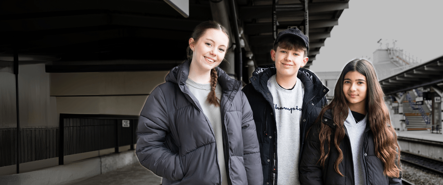 Three teenagers, two girls and one boy (centre), stood at a train station platform smiling at the camera.