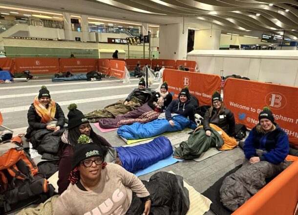 Group of people lay in sleeping bags, wearing bobble hats on Birmingham New Street train station floor.