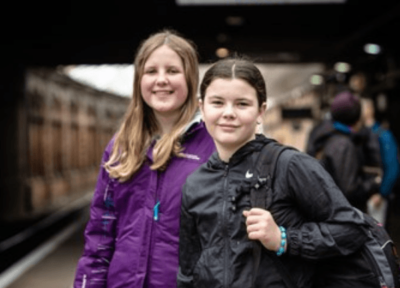Two young girls wearing waterproof jackets at a train station.