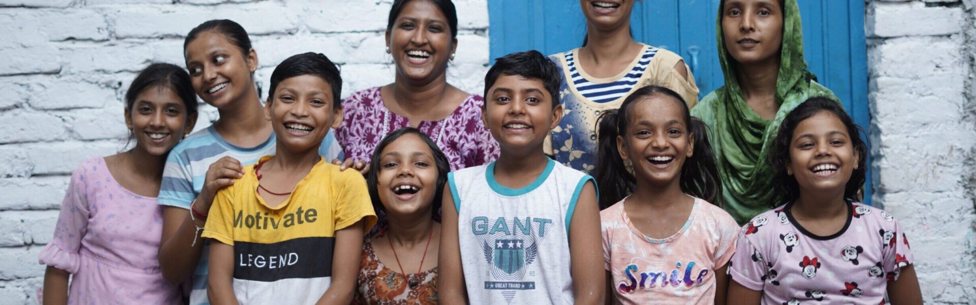 A group of children in India with Railway Children project workers smiling and stood in front of a white wall with a sign that reads 'Children's Activity Centre' next to a blue door.