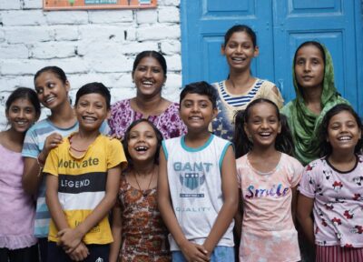 A group of children in India with Railway Children project workers smiling and stood in front of a white wall with a sign that reads 'Children's Activity Centre' next to a blue door.