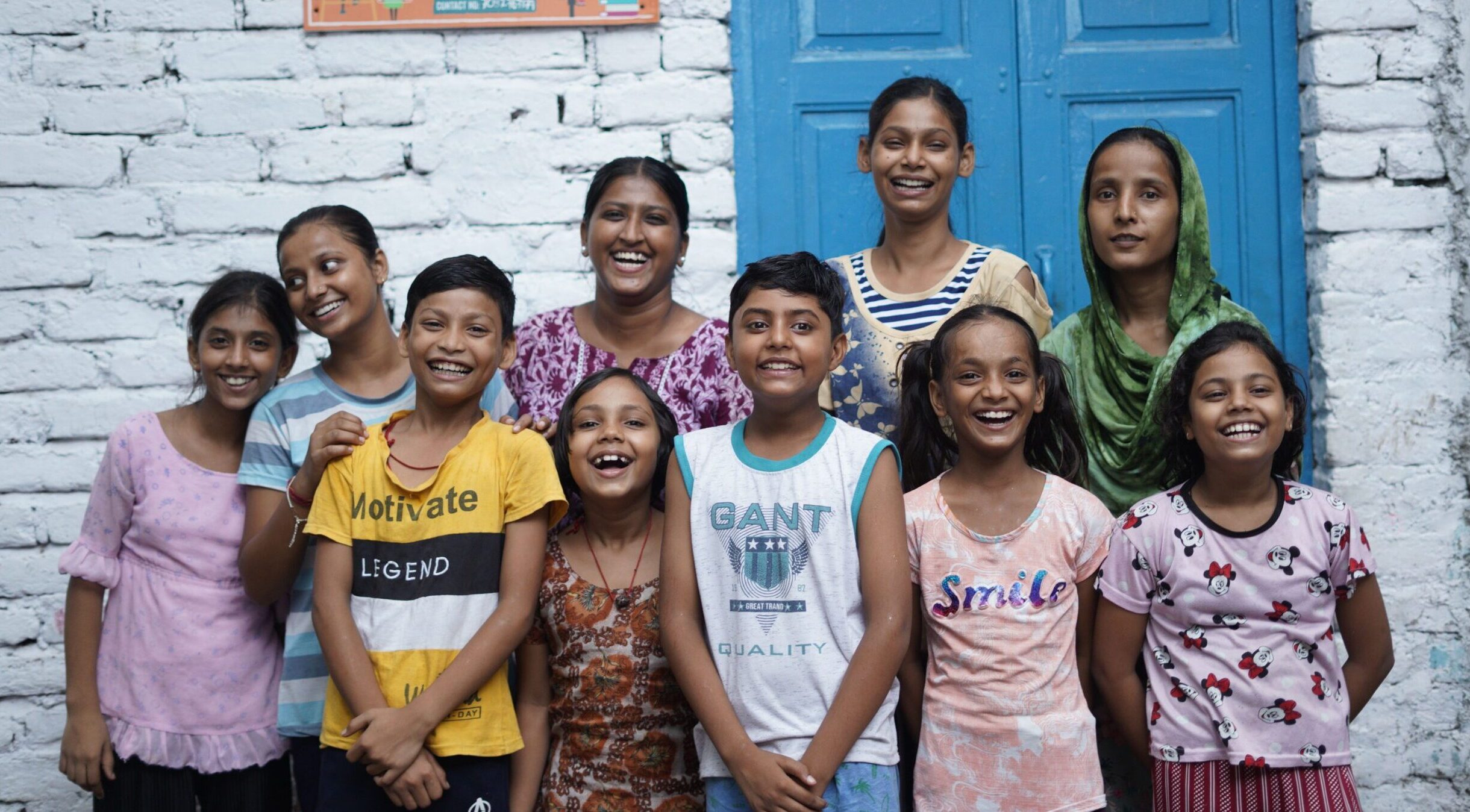 A group of children in India with Railway Children project workers smiling and stood in front of a white wall with a sign that reads 'Children's Activity Centre' next to a blue door.