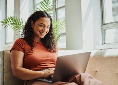 A woman sat on a sofa looking down at her laptop and smiling.