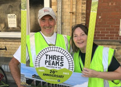 Two Railway Children volunteers wearing hi-vis jackets and holding a Three Peaks By Rail selfie frame.