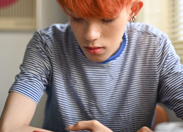 A LGBTQ+ teenager with orange hair wearing a blue striped T-shirt sat leaning on a desk looking down at their phone.