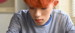 A LGBTQ+ teenager with orange hair wearing a blue striped T-shirt sat leaning on a desk looking down at their phone.