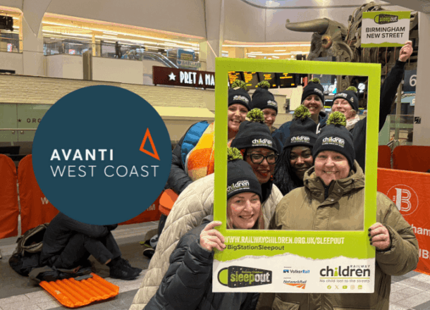 A group of Sleepout participants from Avanti West Coast wearing Railway Children black bobble hats and stood behind a green selfie frame at Birmingham New Street station. Avanti West Coast logo added next to group on a blue circle.