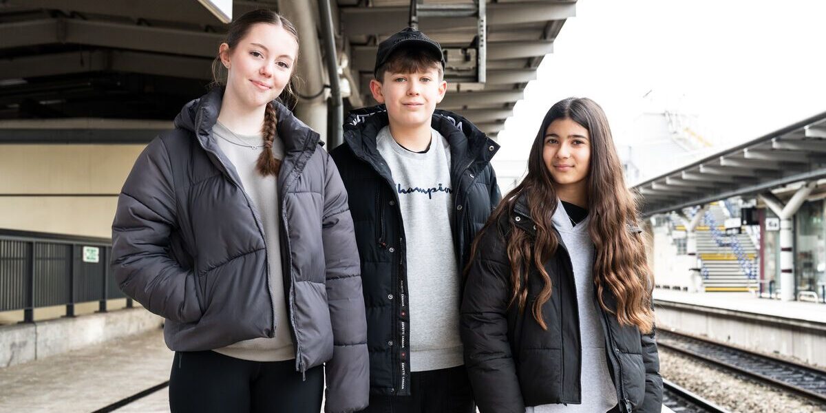 Three teenagers stood smiling for the camera in a train station.