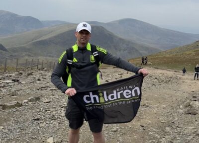 A man in activewear on Mount Snowdon holding a Railway Children flag.