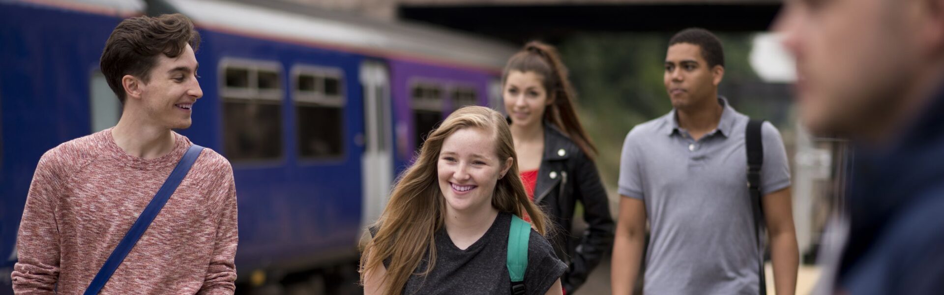 A group of teenagers laughing and smiling walking past a train on a station platform on the UK.