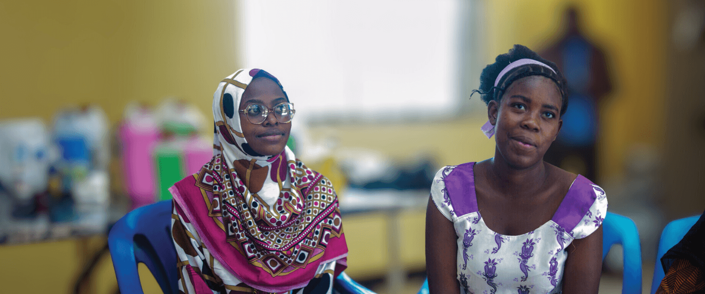 Two teenage girls sat in a classroom on blue chairs in Tanzania.