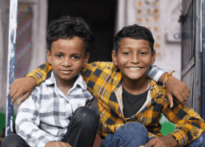 Two boys sat in a classroom smiling with their arms round each other.