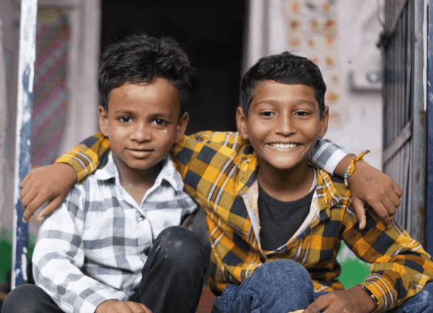 Two boys sat in a classroom smiling with their arms round each other.