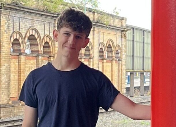 A teenage boy with brown curly hair wearing a navy blue top and leaning against a post at a railway station.