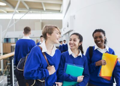 A group of teenage girls in blue school uniform holding books and walking through a school corridor, smiling at each other and chatting.