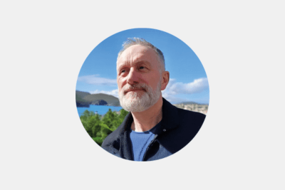 An image of a man in a circle frame on a grey background with grey short hair and a grey beard wearing a blue T-shirt and navy jacket in front of a blue sky and picturesque lake.
